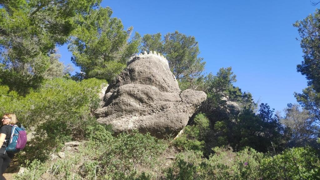 Mirador de sa Panada, con un gran bloque de roca y vegetación densa alrededor. Una persona camina por el sendero en primer plano, bajo un cielo despejado.