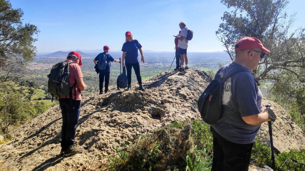 Mirador de la Panda y Penyal de los Bous - Caminando por Mallorca Penyal dels Bous, con vistas panorámicas del paisaje montañoso y campos abajo. Algunos miembros del grupo están de pie y otros en una posición más baja, todos usando gorras rojas y mochilas.