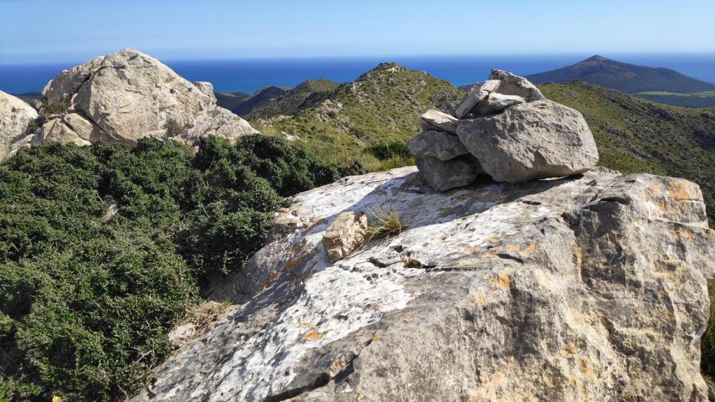 Vista panorámica desde la cima de Puig Poloni, ,en la ruta 'Serra de Sos Sastres', con rocas y vegetación en primer plano y el mar al fondo.