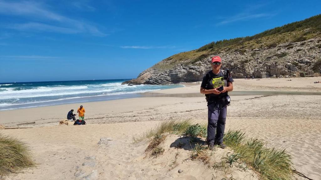 Persona de pie en la playa de Cala Torta, con suéter negro y gorra roja, sosteniendo una cámara. Al fondo, se observa la playa con olas y una colina. Dos personas sentadas en la arena a la izquierda.