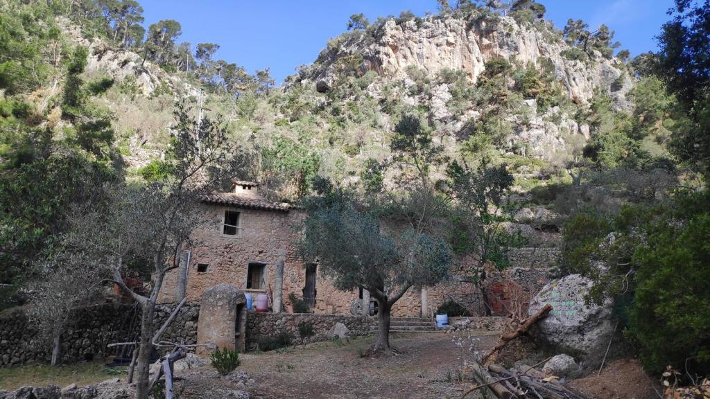 Vista de una casa rústica de piedra rodeada de vegetación en un entorno montañoso, con un acantilado al fondo.