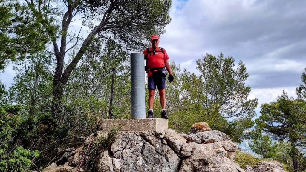 Hombre de pie sobre una base rocoso con un vértice geodésico, rodeado de árboles y vegetación en un paisaje montañoso.