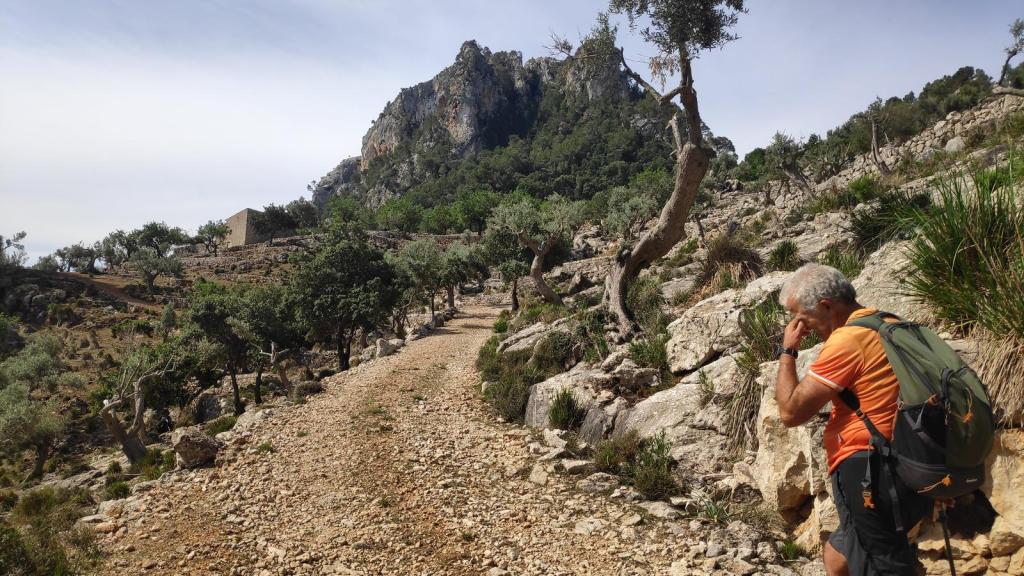 Hombre en una ruta de montaña con un paisaje de rocas y árboles, mirando pensativo hacia el Puig de n'Escuder.