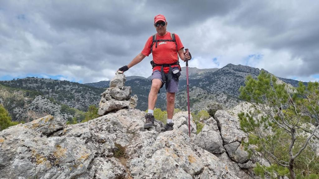 Hombre de pie sobre una roca en la cima del Puig de n’Escuder, vestido con camiseta roja y pantalones cortos, sosteniendo un bastón de senderismo, rodeado de un paisaje montañoso y nubes en el cielo.