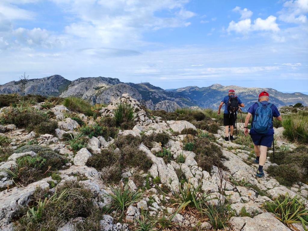 Vista de excursionistas caminando por un sendero de montaña rodeado de rocas y vegetación, con montañas al fondo bajo un cielo parcialmente nublado.