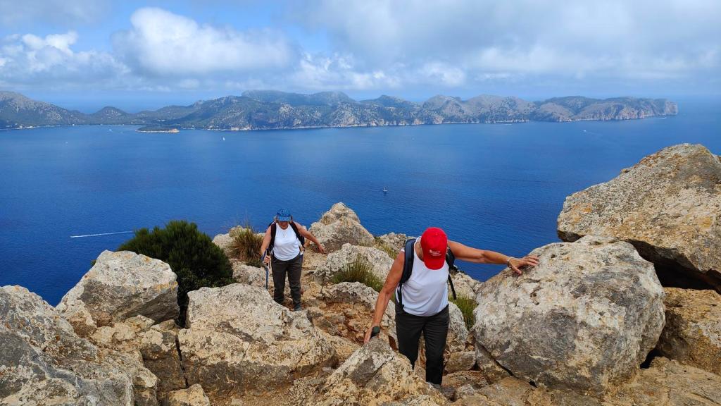 Personas haciendo senderismo en una cima montañosa, con vistas panorámicas del mar y las montañas en el fondo.
