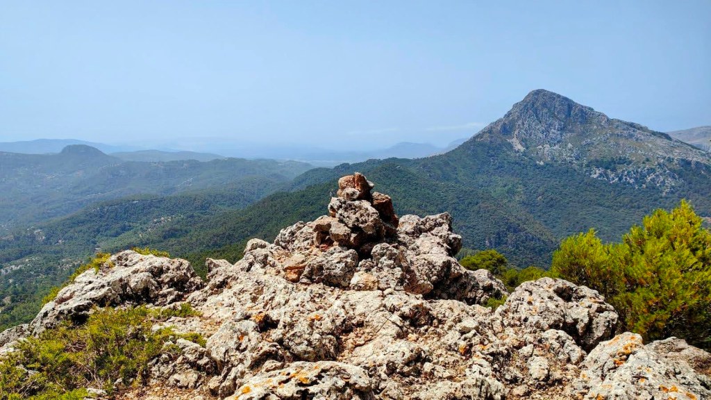 Vista desde un pico montañoso en la Serra de Tramuntana, con un fondo de montañas verdes y un gran pico rocoso al fondo.