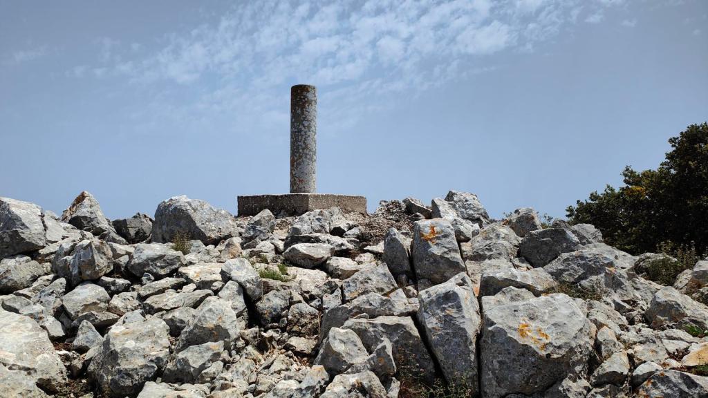 Punto geodésico en la cima de una montaña rodeado de rocas y vegetación en el fondo con un cielo parcialmente nublado.