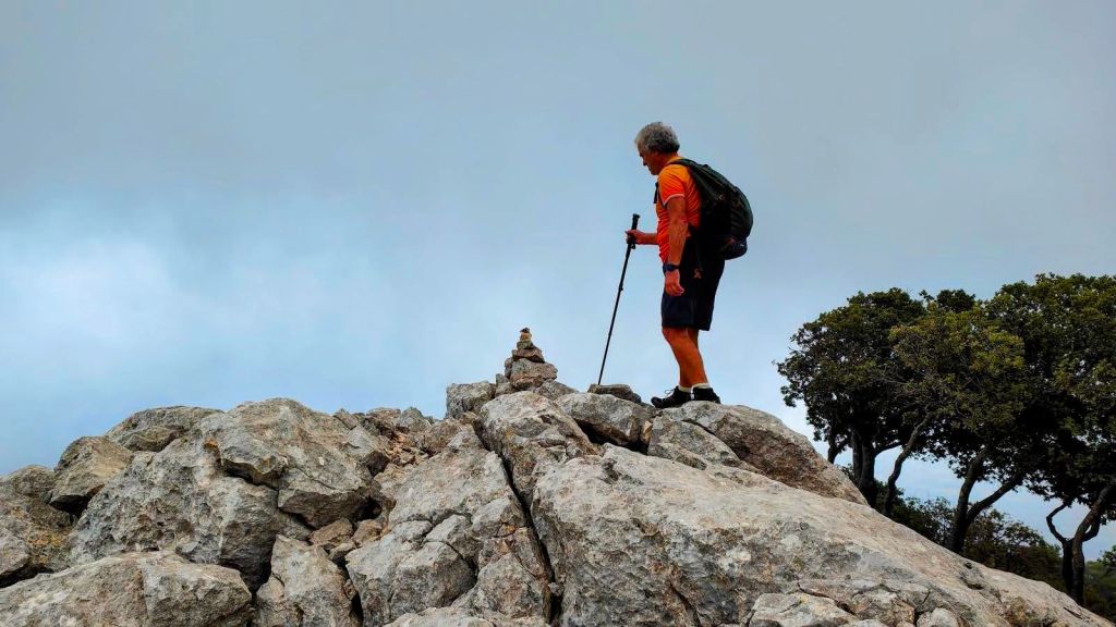 Fita del Ram por el Pas de Gotleu - Caminando por Mallorca Un senderista de pie sobre la cima de Na Ferrana mirando hacia un hito o señal de montaña, con un fondo nublado.