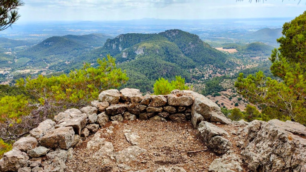 Fita del Ram por el Pas de Gotleu - Caminando por Mallorca Vista panorámica desde un mirador natural, con un frente de montaña verde y una amplia llanura hacia el horizonte.