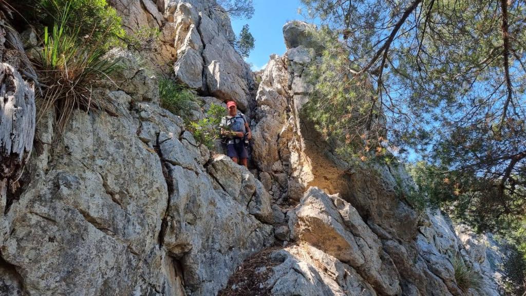 Hombre escalando el  Pas de sa Servera, una roca en un sendero de montaña con vegetación alrededor, bajo un cielo azul.