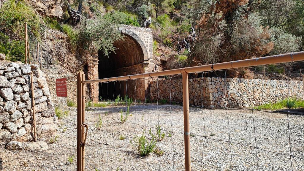 Entrada de un túnel de piedra en un camino de montaña rodeado de vegetación. Se observa una valla metálica en primer plano junto a un rótulo informativo.