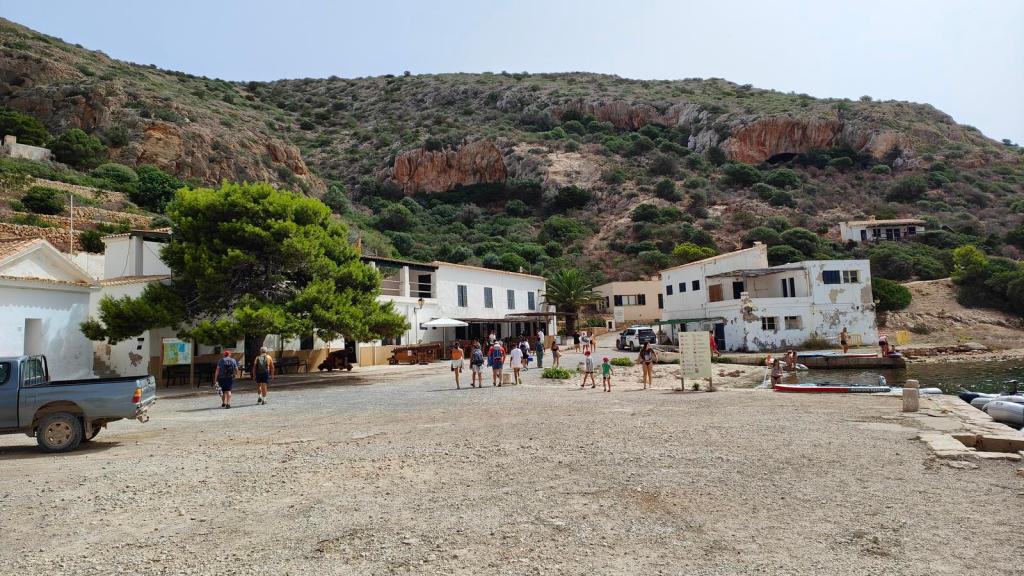 Vista del Puerto de Cabrera, con edificios de servicios y visitantes en la costa junto al mar, rodeado de colinas verdes.