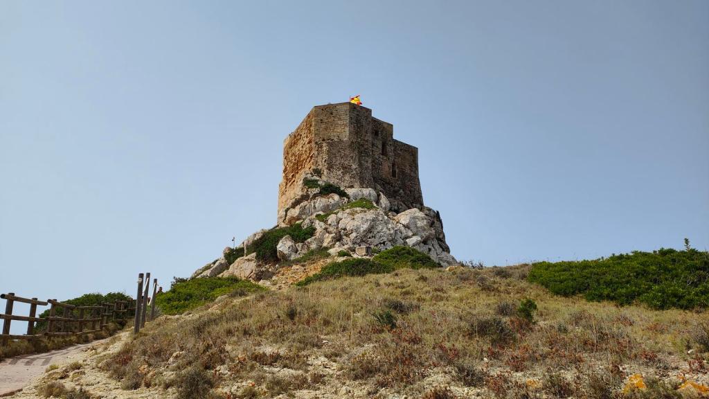 Vista del Castillo de Cabrera, una edificación histórica situada en un acantilado con vegetación circundante y una bandera ondeando en la cima.