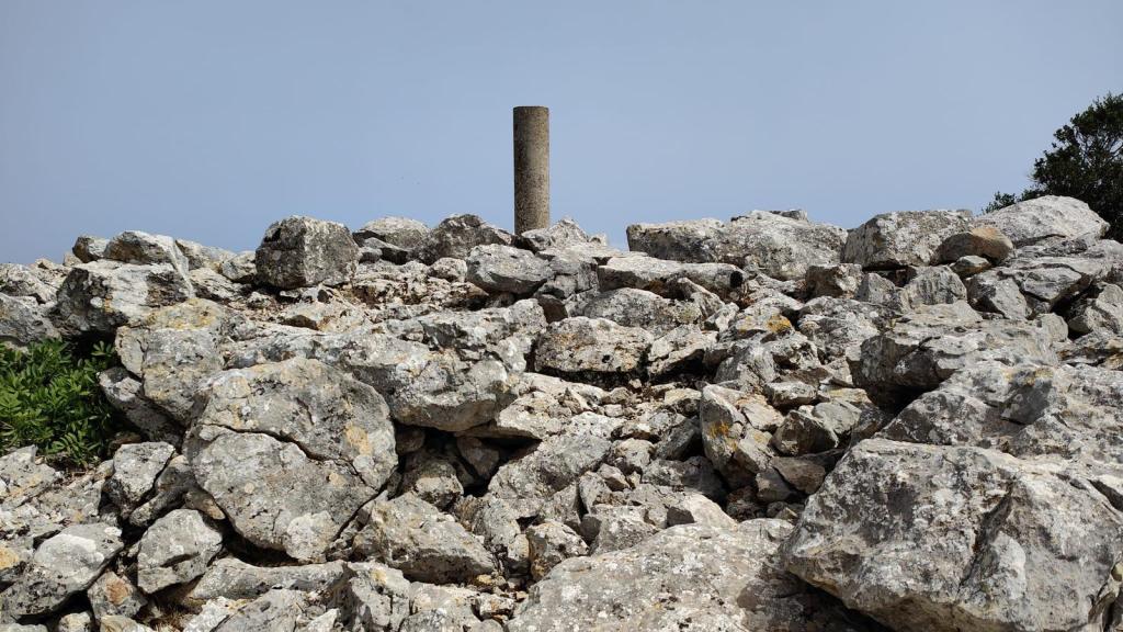 Vista de un terreno rocoso en la cima de la Peña Mascorda, con un monolito en pie sobre una superficie de piedras. El cielo está parcialmente nublado.