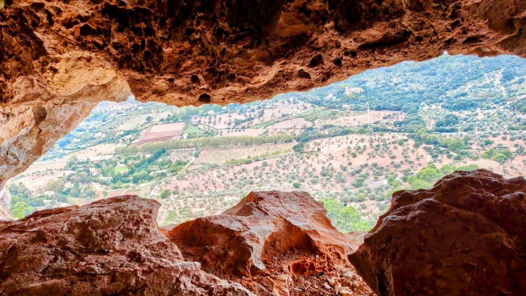 Cueva de las dos Bocas-Pas de Son Moragues - Caminando por Mallorca Vista desde el interior de la Cova de ses Dues Boques, con rocas en primer plano y un paisaje agrícola y montañoso al fondo.
