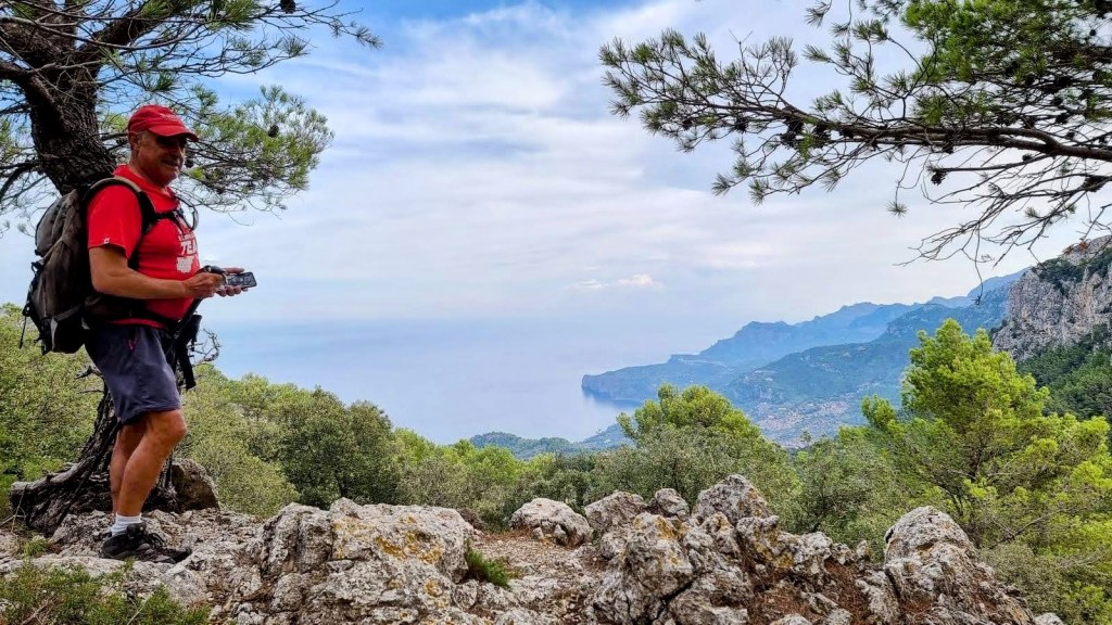Cueva de las dos Bocas-Pas de Son Moragues - Caminando por Mallorca Hombre con mochila y gorra roja, sosteniendo una cámara, posando en un paisaje montañoso con vistas al mar y árboles alrededor.