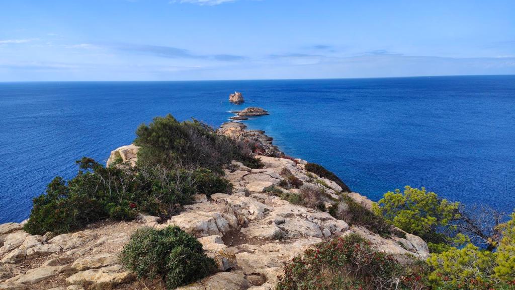 Punta del Toro y Cala Rafeubetx - Caminando por Mallorca Vista panorámica de acantilados y mar en la ruta Punta del Toro y Cala Rafeubetx, con vegetación costera y un islote visible en el horizonte.