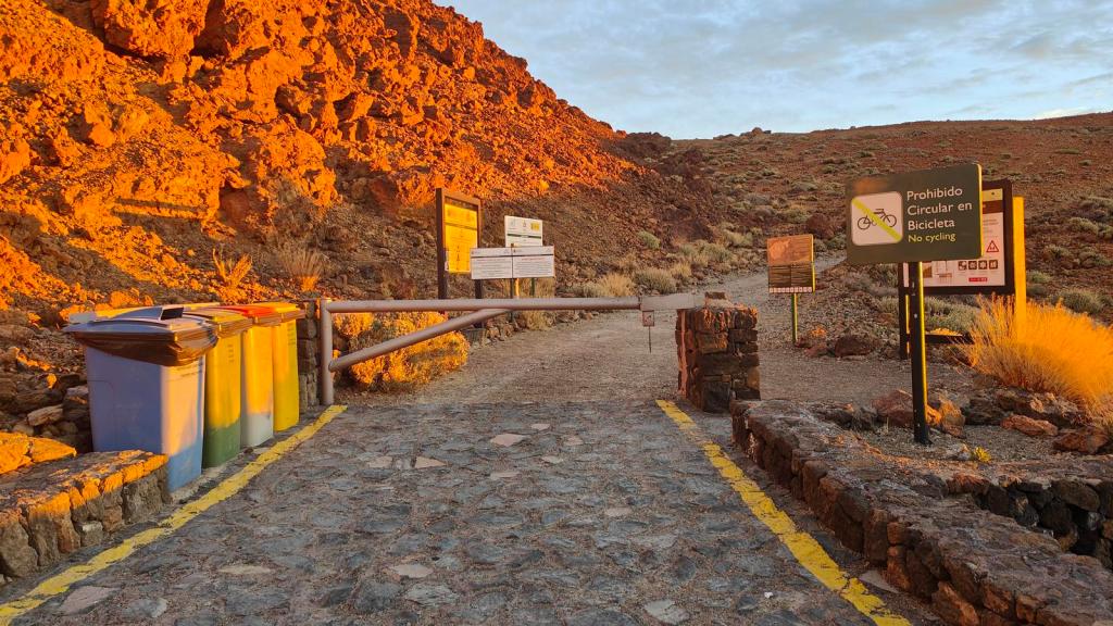 Entrada al sendero de Montaña Blanca, con señales de advertencia y contenedores de reciclaje en un paisaje volcánico iluminado por el sol de la mañana.