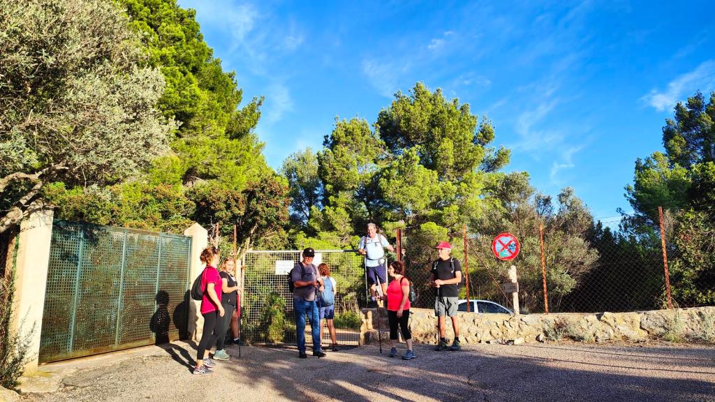 Grupo de excursionistas reunidos frente a una puerta en el inicio de la ruta 'Coll de Cala Ferrera desde el Port de Sóller', rodeados de vegetación y con un cielo azul claro.