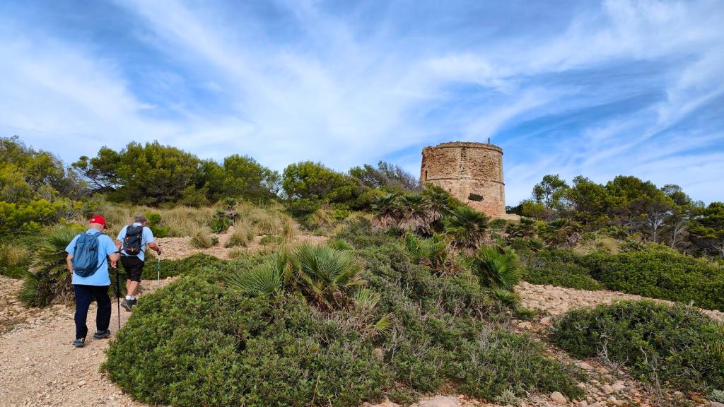 Dos senderistas subiendo por un camino natural hacia la Torre d'Albarca, rodeados de vegetación y cielo parcialmente nublado.