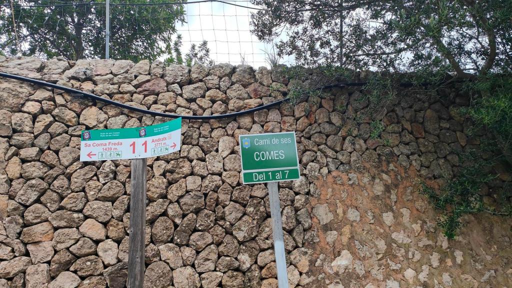 Señales direccionales de la Ruta de ses Comes d'Andratx, con instrucciones hacia Sa Coma Freda y Andratx, frente a un muro de piedra.
