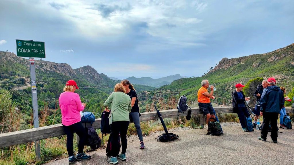 Grupo de personas en una parada durante la ruta de senderismo, junto a un cartel que indica 'Camí de sa Coma Freda', con montañas y paisaje verde de fondo.