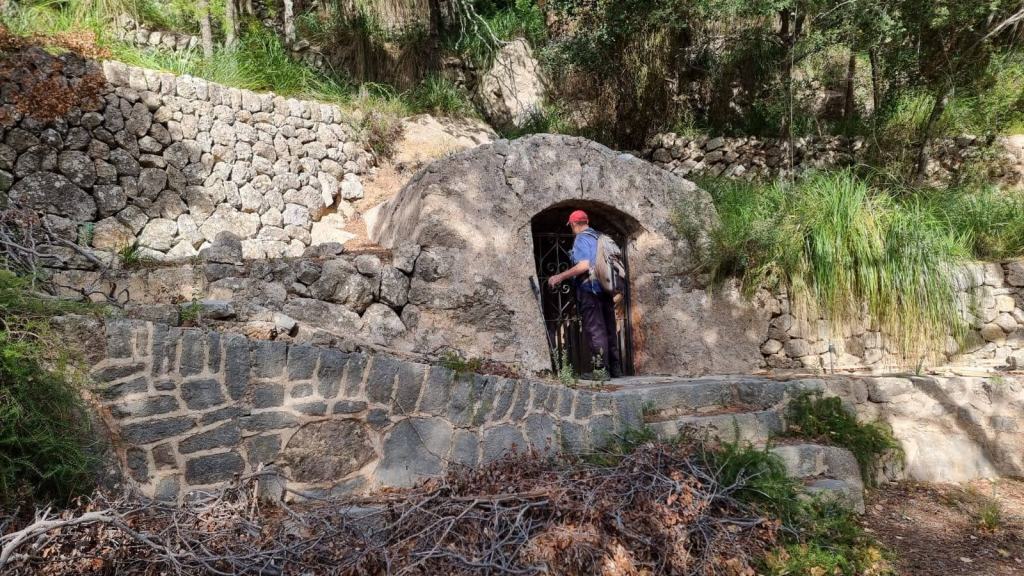 Hombre con mochila acercándose a una puerta de metal en una estructura de piedra rodeada de vegetación.