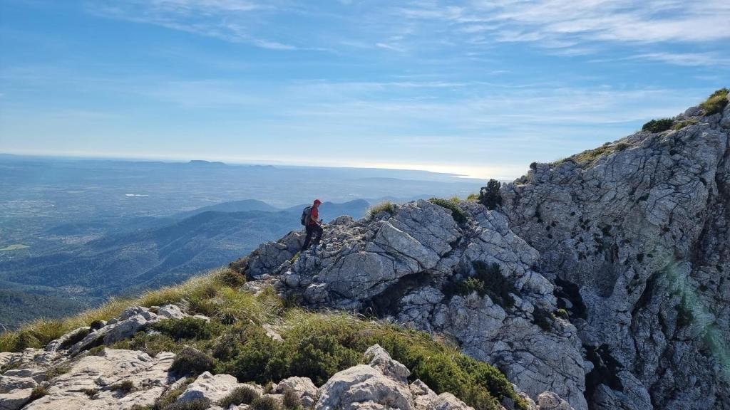 Persona caminando sobre rocas en la cima de una montaña, con un paisaje montañoso y un cielo despejado de fondo.