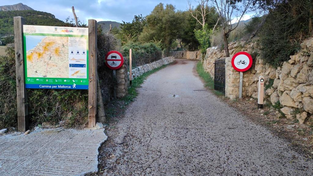 Señalización de la ruta 'Camina per Mallorca' con un mapa y restricciones de acceso, incluyendo letreros que prohíben vehículos y bicicletas, en un camino rodeado de vegetación y muros de piedra.