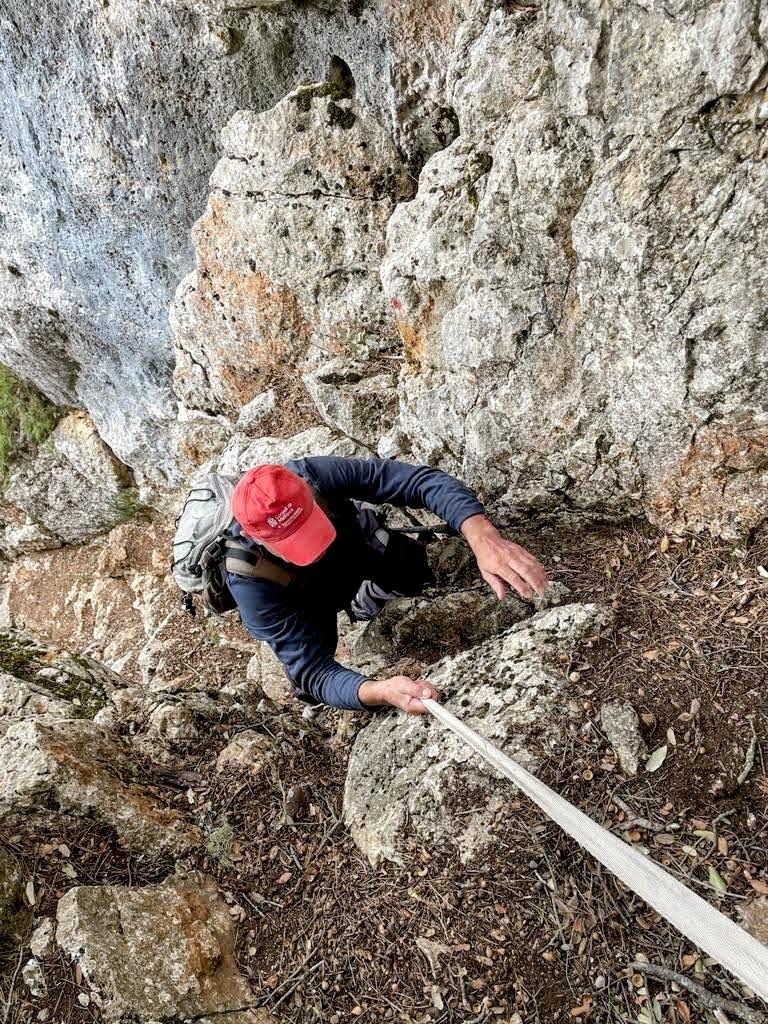 Hombre escalando una roca inclinada con una cuerda, en un entorno natural rocoso.