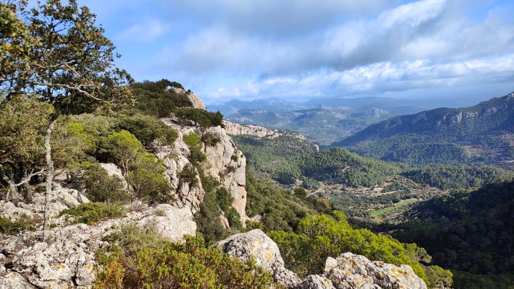Vista panorámica desde el sendero en la sierra de Puntals de Planicia, con rocas y vegetación en primer plano y un paisaje montañoso al fondo bajo un cielo nublado.