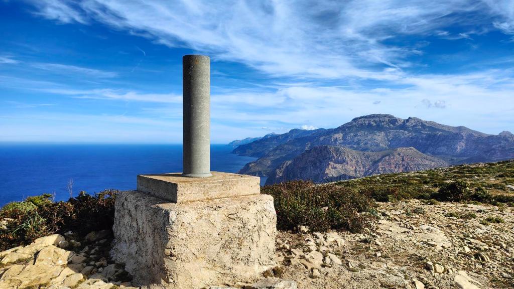 Vértice geodésico en la cima del Puig de ses Basses, con vistas al mar y montañas de fondo bajo un cielo azul.