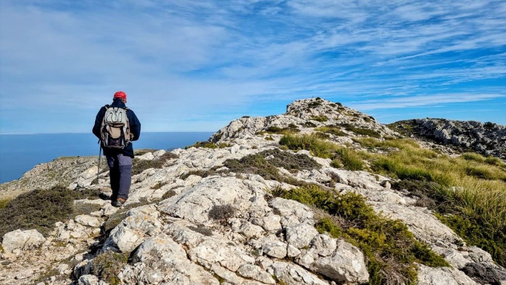 Una persona camina por un sendero rocoso en la cima de una montaña, con vistas al mar y cielo despejado al fondo.