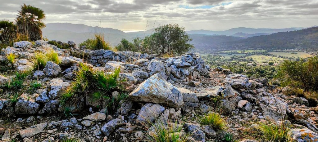 Vista desde una cima montañosa con restos de piedras y vegetación, en el fondo se aprecian montañas y un paisaje natural.