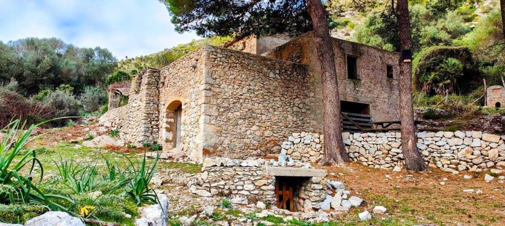 Vista de un refugio de piedra rodeado de vegetación en un entorno natural, con árboles y piedras en el primer plano.