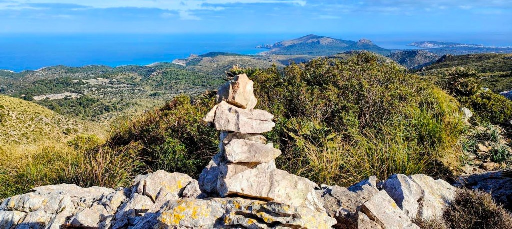 Vista panorámica desde la cima de Sa Moleta con un hito de piedras apiladas en primer plano, rodeado de vegetación y con el mar y las montañas al fondo.