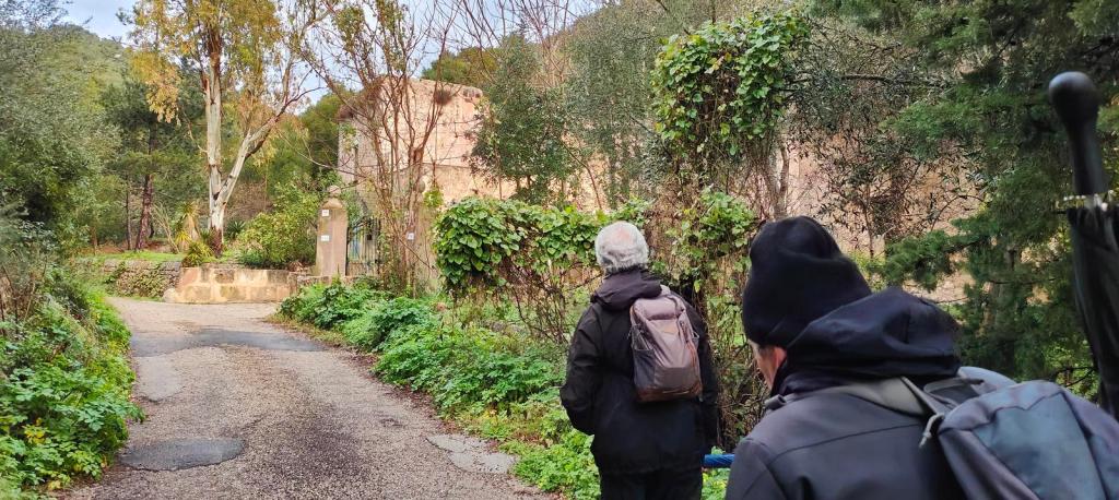 Dos excursionistas recorren un camino rodeado de vegetación en dirección a las casas de sa Font des Jardí, en un sendero montañoso.