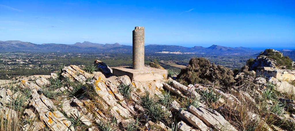 Vista panorámica desde la cima de la Torreta des Tressor,, con un hito geodésico en primer plano, rodeado de rocas y vegetación, y un paisaje montañoso al fondo.