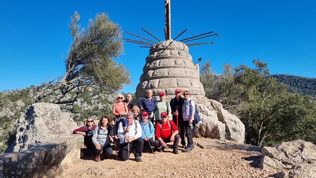 Grupo de excursionistas posando junto a un hito con una cruz en la cima del Pujolet des Misteris, con un paisaje montañoso al fondo y un cielo despejado.