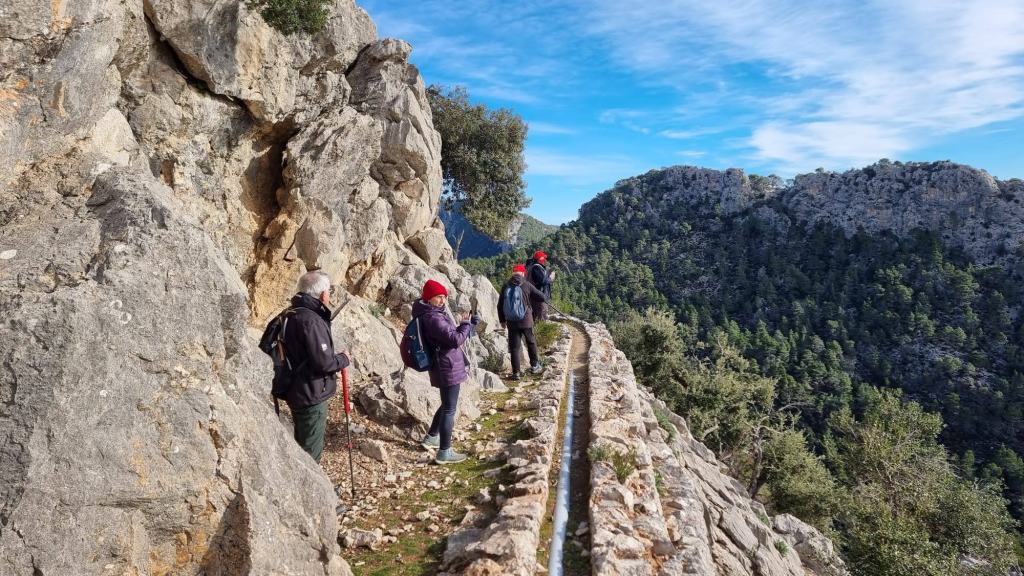 Grupo de senderistas caminando por un sendero estrecho junto a una canaleta en una zona montañosa, rodeados de rocas y vegetación.