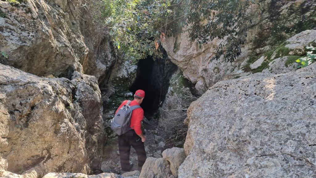 Es Pixarells desde Lluc - Caminando por Mallorca Hombre de pie frente a la entrada de una cueva, rodeado de rocas y vegetación, mientras explora el entorno natural.
