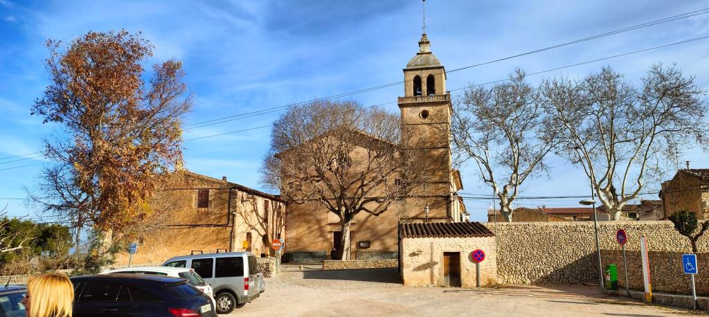 Vista del aparcamiento frente a la iglesia dedicada al beato Ramón Llull en Randa, Mallorca. Se observa un edificio con campanario, árboles sin hojas y vehículos aparcados.