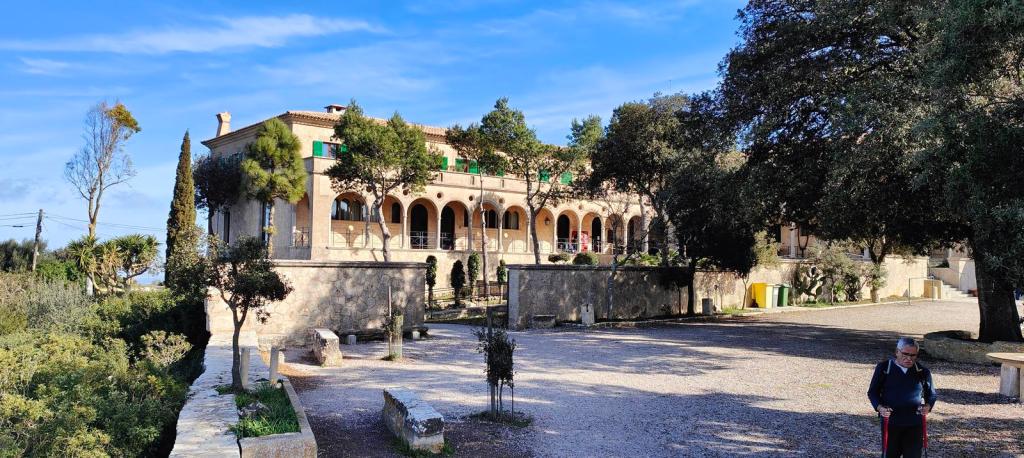 Vista del Santuario de Cura, un edificio con arcos y terraza, rodeado de vegetación en el Puig de Randa, Mallorca. Se observa un camino de acceso y un visitante con bastón en primer plano.