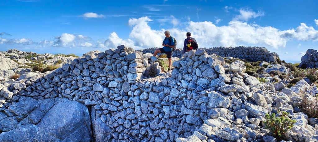 Es Frontó de Comafreda - Caminando por Mallorca Dos personas de pie sobre una construcción de piedra en un paisaje montañoso bajo un cielo azul con algunas nubes.