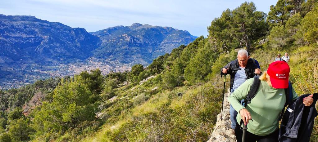 Senderistas ascendiendo por un sendero montañoso con vistas panorámicas de la Serra de Tramuntana y el Valle de Sóller. La vegetación es densa y el paisaje es impresionante.