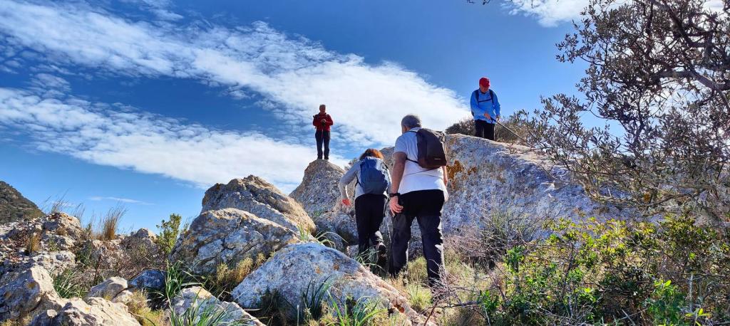 Grupo de excursionistas ascendiendo por un terreno rocoso en la ruta Puig de ses Bruixes-Puig de s'Escolà, con un cielo azul y algunas nubes.