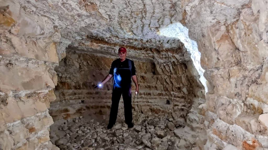 Un hombre de pie en el interior de una cueva iluminando el espacio con una linterna, rodeado de paredes de piedra y suelo rocoso.