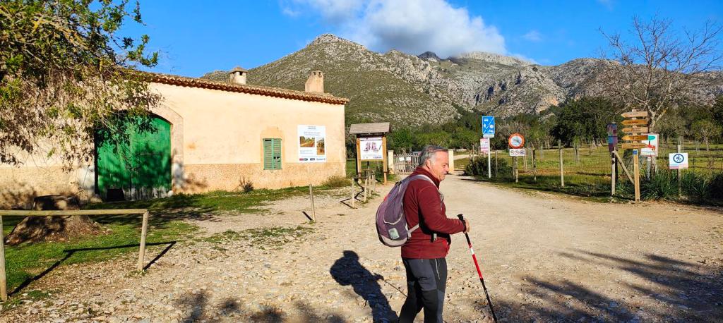 Un senderista con bastón camina por un camino terrado junto a una casa de campo con puerta verde, con montañas al fondo bajo un cielo azul.