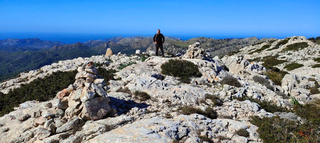Puig Batiat-Cadira del Bisbe-Es Moletó - Caminando por Mallorca Vista panorámica desde la cima de una montaña, con un hombre en pie junto a un hito de piedras, rodeado de un paisaje rocoso y montañoso, con el mar al fondo y un cielo despejado.